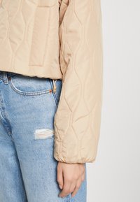 Beżowa pikowana kurtka z teksturowanym, sześciokątnym wzorem, krótki fason i boczna kieszeń, noszona na jasnoniebieskich, postarzałych jeansach.