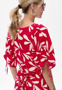 Frau mit blonden Pferdeschwanz, die große Hoop-Ohrringe und ein rotes Kleid mit weißen, abstrakten Blattmustern und gerafften Ärmeln trägt.