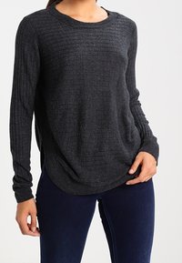 Frau trägt dunkelgrauen, gerippten Pullover mit langen Ärmeln und dunkelblaue Jeans, steht mit einer Hand in der Hüfte und der anderen locker.