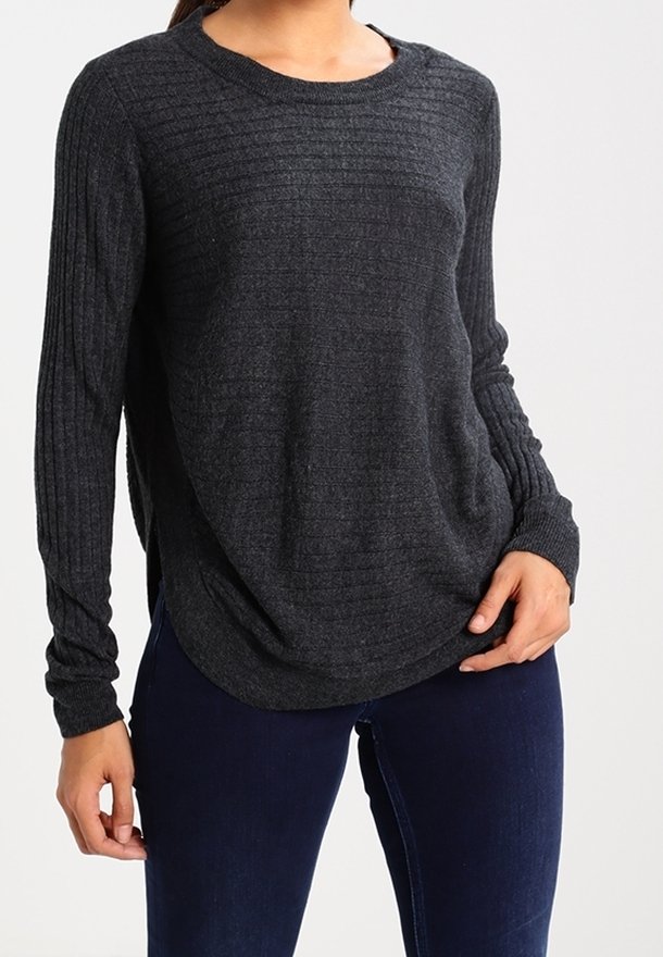 Frau trägt dunkelgrauen, gerippten Pullover mit langen Ärmeln und dunkelblaue Jeans, steht mit einer Hand in der Hüfte und der anderen locker.