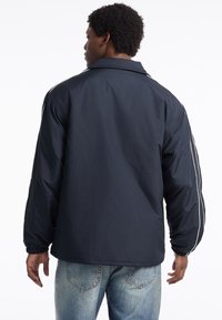 Marineblaue Windjacke aus leichtem Material, mit lockerer Passform, weißen seitlichen Streifen und elastischen Bündchen, versehen mit einem Rückenschlitzdetail.