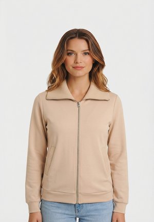 Femme aux cheveux châtain clair portant une veste beige zippée et un jean bleu clair, faisant face à l'avant avec une expression neutre.