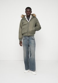 Olive rohelise bomber-jakk, mille kapuuts on kaetud karvaga, lukuga kinnitatav, kantud halli särke ja siniseid laia säärega teksaseid peal, valgete nokatsitega.