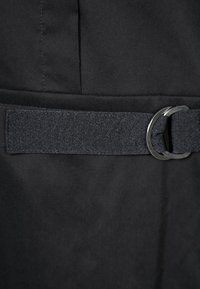 Ceinture en tissu noir avec une lanière texturée passant à travers deux anneaux métalliques ronds sur un vêtement foncé.
