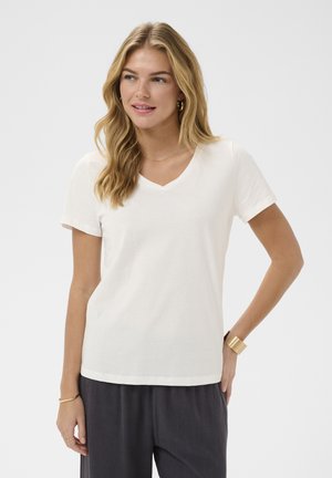 Donna con capelli biondi lunghi che indossa una maglietta bianca a V, pantaloni scuri, orecchini a cerchio dorati, braccialetto e polsino, che guarda verso destra.