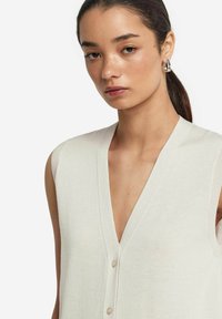 Ljust beige ärmlös cardigan med djup V-hals, knappstängning, ribbad textur och smidig tyg. Nätt avslutade kanter.