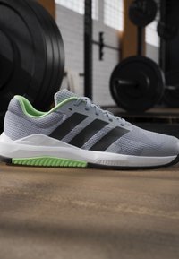 Chaussure de sport grise et noire avec intérieur vert et détail de la semelle, posée sur le sol de la salle de sport avec du matériel de musculation en arrière-plan.