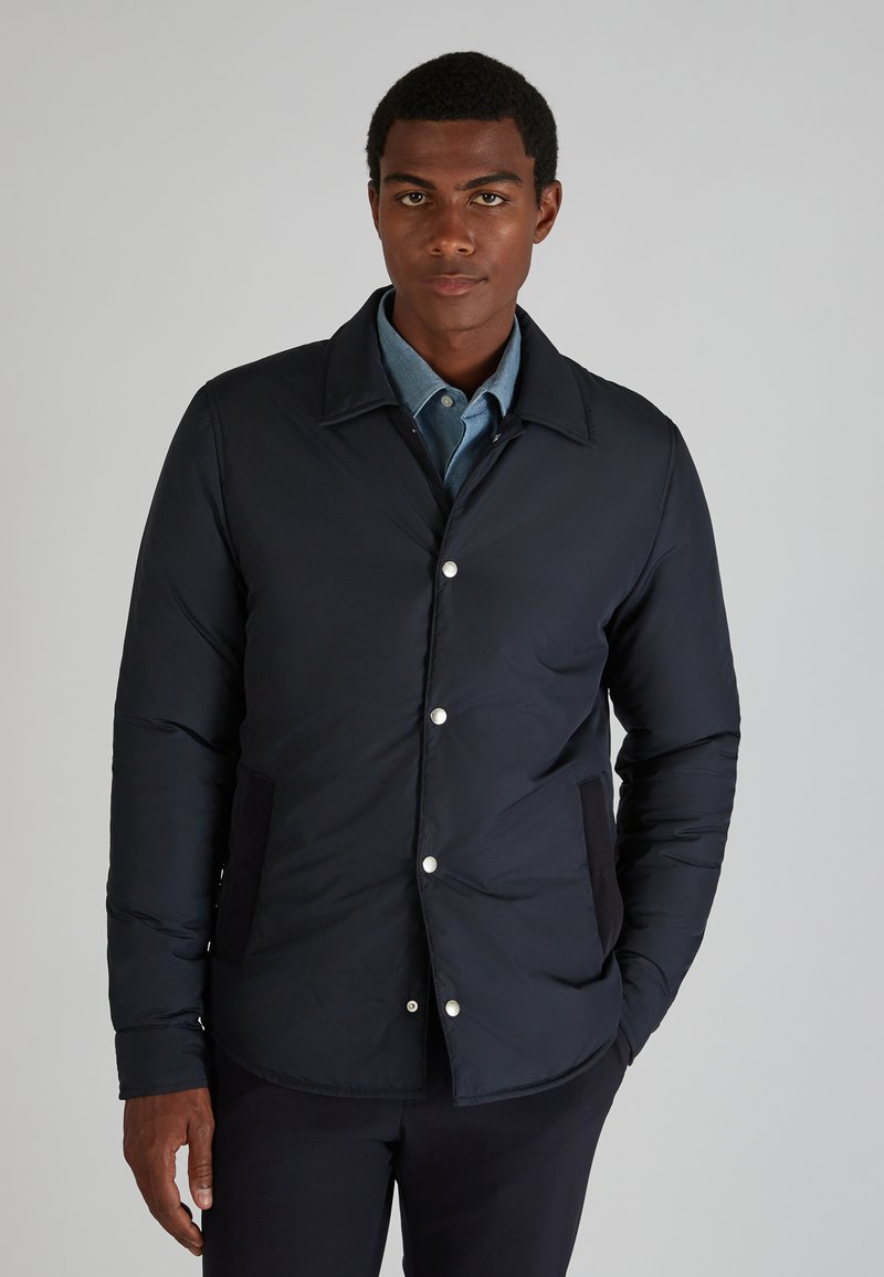 Giubbotto imbottito navy con design colletto, bottoni a pressione argentati e tasche laterali. Tessuto liscio con una leggera texture trapuntata.