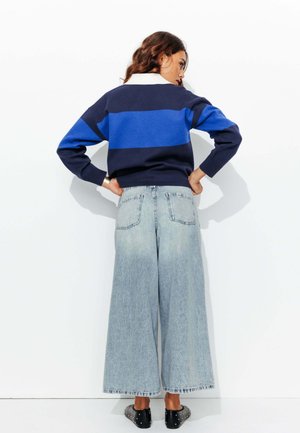 Pull rayé bleu marine et bleu avec un col blanc, associé à un jean en denim bleu clair à jambe large. Chaussures noires avec des clous argentés.