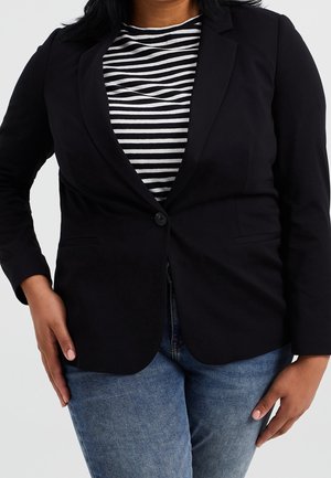 Persona che indossa un blazer nero con un solo bottone sopra una maglietta a righe bianche e nere con jeans blu, in piedi con le mani rilassate lungo i fianchi.