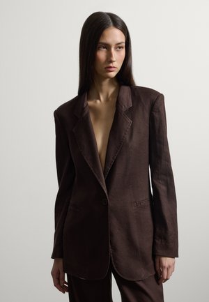 Frau mit langen dunklen Haaren, die einen übergroßen dunkelbraunen Blazer und passende Hosen trägt, steht vor einem schlichten hellen Hintergrund.