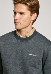 Sudadera gris de tela suave, con cuello redondo y logo bordado de "PEPE JEANS" en el pecho, combinada con una camisa de cuadros.