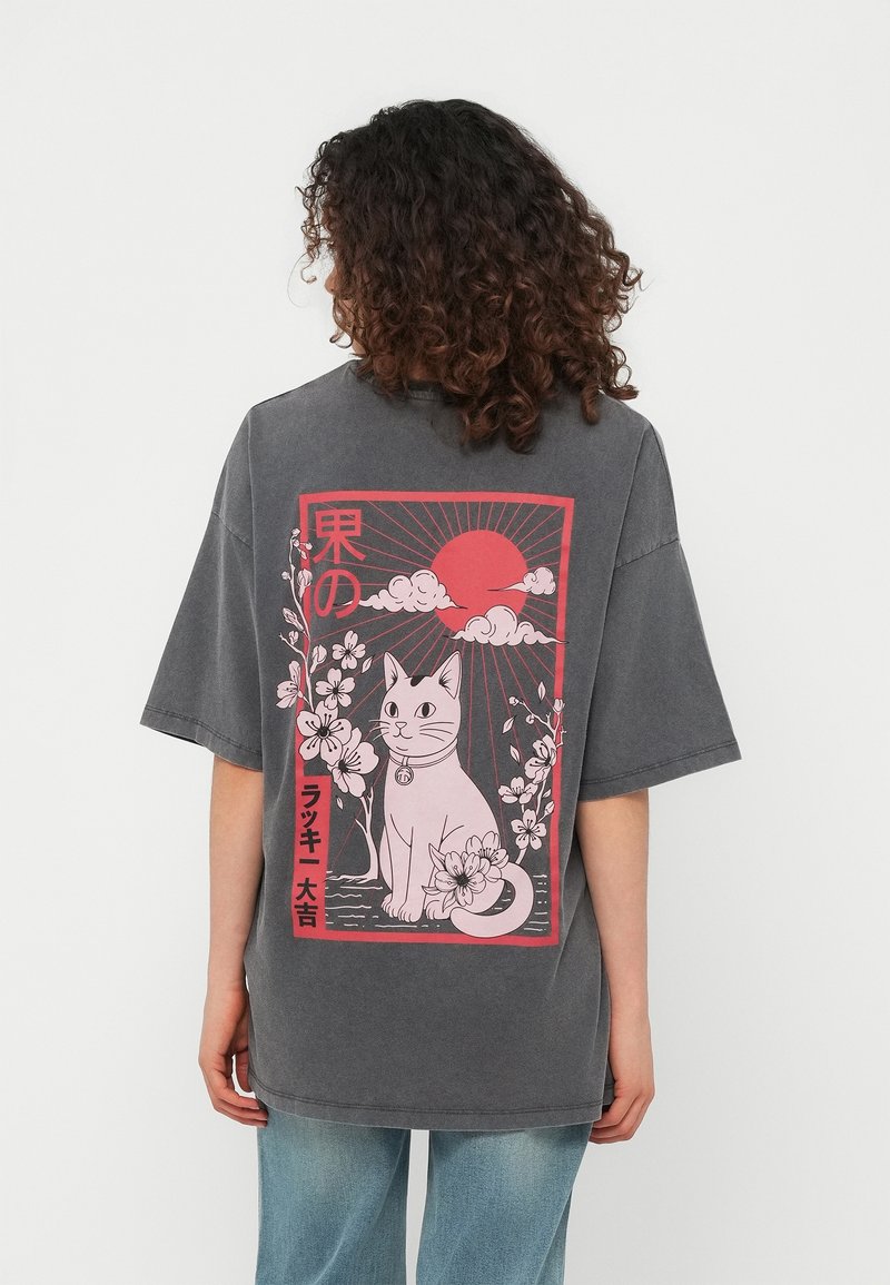 Personne aux cheveux bouclés portant un t-shirt gris avec un design de chat rose et floral avec soleil et nuages dans le dos.