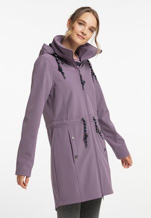 Junge Frau trägt eine lange violette Kapuzenjacke mit schwarzen Kordeln, steht vor einem schlichten weißen Hintergrund.