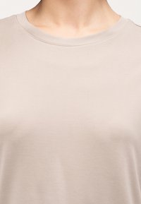 Κοντινό πλάνο ενός ατόμου που φοράει απλό μπεζ t-shirt με στρογγυλή λαιμόκοψη, εμφανίζοντας την περιοχή του λαιμού και του άνω στήθους σε λευκό φόντο.