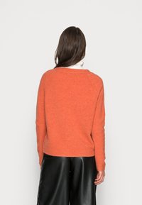 Kvinna med långt brunt hår som bär en orange tröja över en vit polotröja och svarta läderbyxor med vida ben, stående vänd bort.