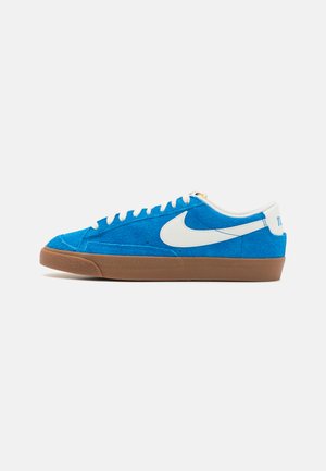 Blå semskede sneakers med en hvit Nike swoosh, brun gummisåle og hvite snører. Har et low-top design og sømdetaljer på overdelen.