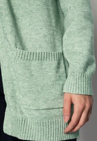 Světlé zelené cardigan z jemně strukturovaného pletení; má žebrované manžety a lem a náprsní kapsu.