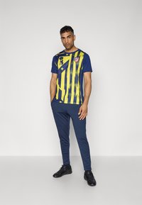 Marineblå og gul sportstrøje med stribet design, logo på brystet og matchende marineblå bukser. Har en kontrasterende tekstur og atletisk pasform.
