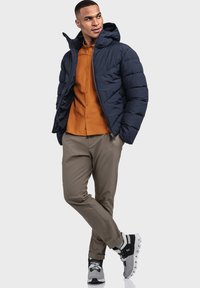 Mann trägt eine navyblaue Daunenjacke über einem orangefarbenen Hemd, beigefarbenen Hosen und schwarz-weißen Sportschuhen und steht mit gekreuzten Beinen.