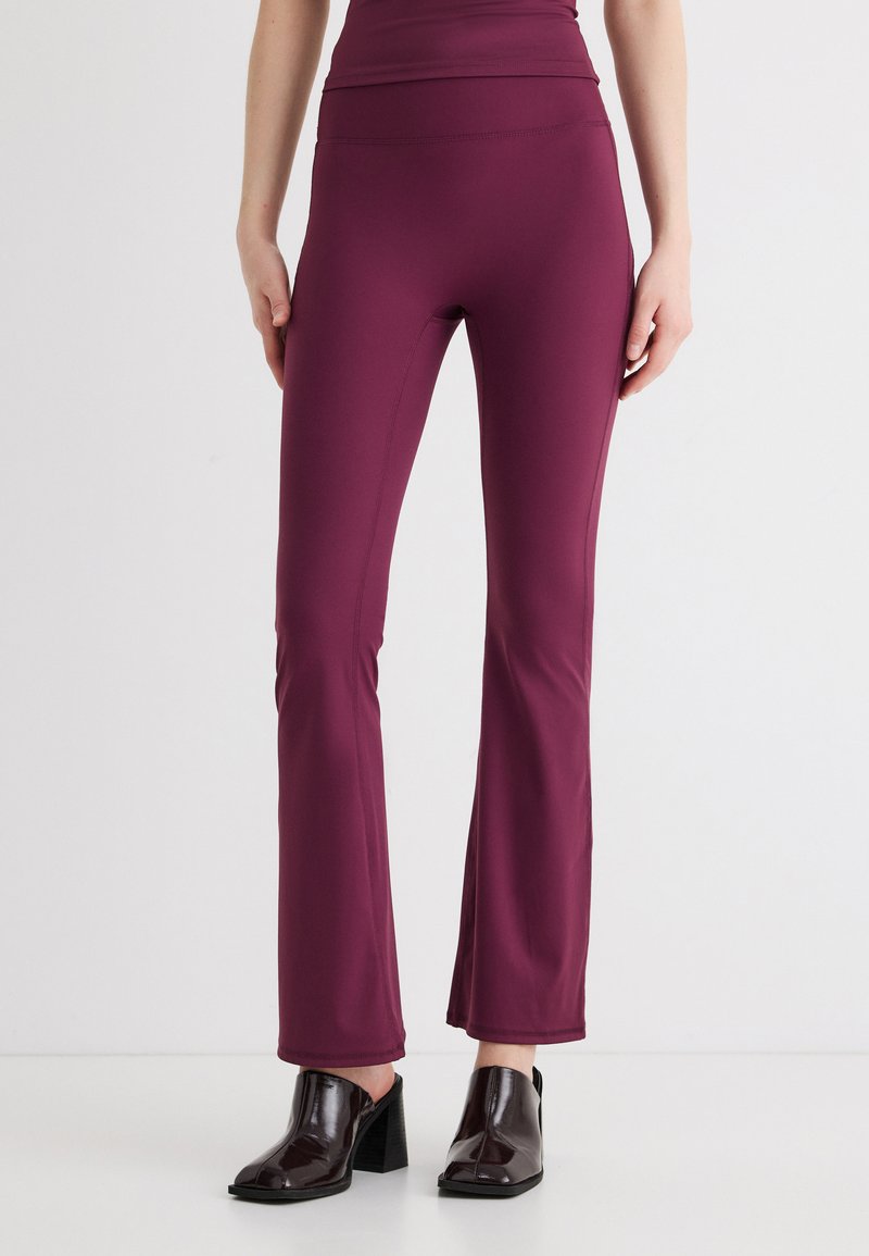 Γυναίκα που φοράει εφαρμοστά καμπανούλα παντελόνια χρώματος δαμάσκηνου και μαύρες μπότες αστραγάλου με τακούνι, πάνω σε απλό ανοιχτό φόντο.