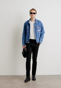 Blauje Denimjacke, weißes T-Shirt, schwarze Hose und schwarze Schuhe. Hält eine schwarze Tasche. Sonnenbrille getragen. Glatte Texturen und taillierter Schnitt.