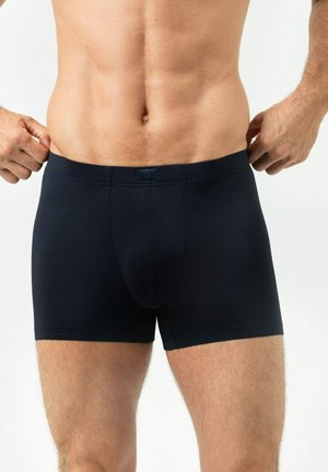 Männlicher Oberkörper und obere Beine in schwarzen, enganliegenden Boxershorts, Hände justieren den Bund vor einfachem hellem Hintergrund.