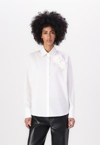 Camicia bianca con colletto strutturato e due fiori di tessuto sul lato sinistro della spalla. Maniche lunghe, vestibilità rilassata, texture liscia.