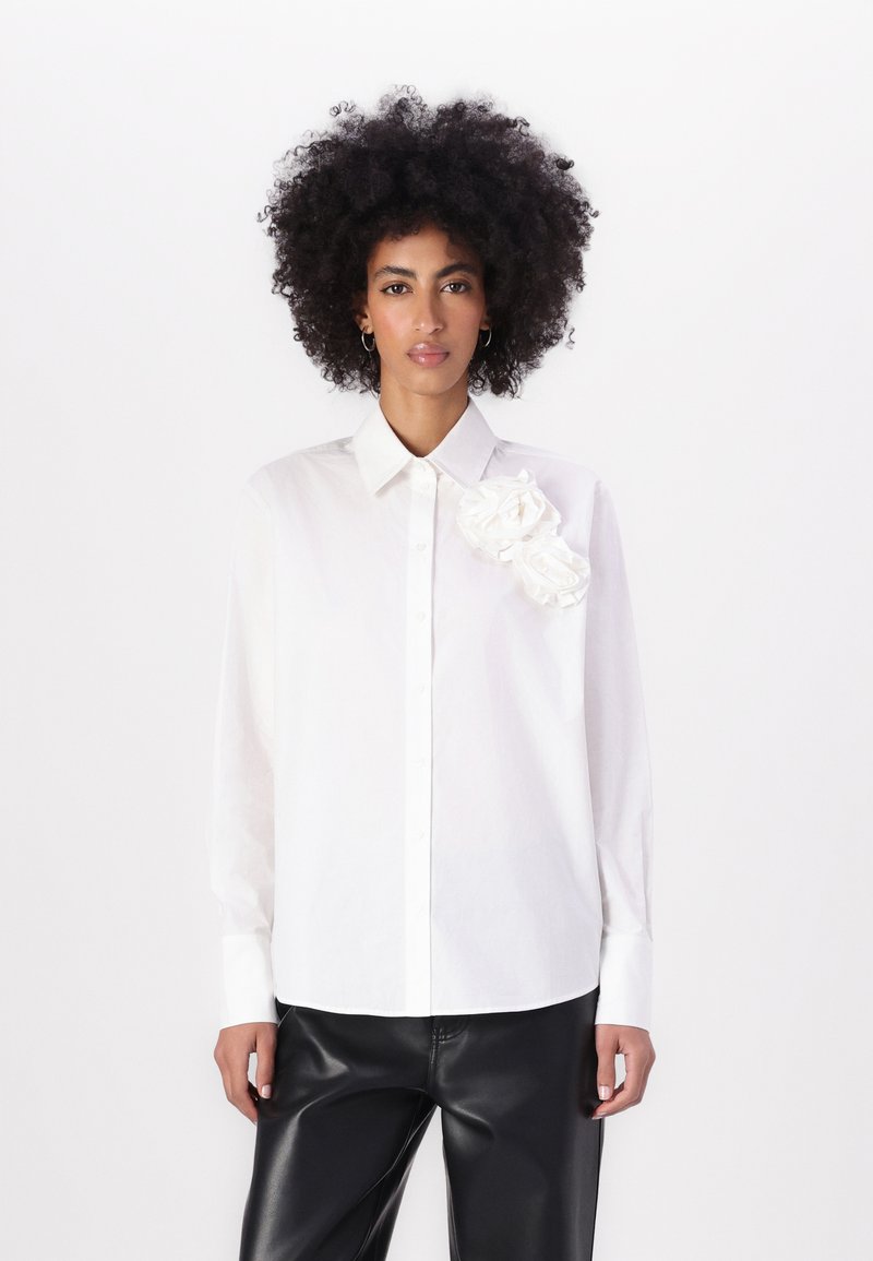 Camicia bianca con colletto strutturato e due fiori di tessuto sul lato sinistro della spalla. Maniche lunghe, vestibilità rilassata, texture liscia.