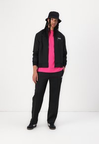 Svart treningjakke, rosa t-skjorte, svarte bukser, og svart bøttehatt med flettet hår. Har subtil merkevare og sporty designelementer.