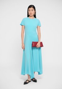 Sinivihreä täysipitkä mekko lyhyillä hihoilla, norjalla helma ja korkealla alhaalla. Malli pitää punaista clutch-laukkua ja hänellä on mustat ballerinat.