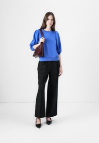 Felpa blu con maniche a sbuffo, abbinata a pantaloni neri a gamba larga e décolleté neri appuntiti, mentre tiene una borsa bordeaux.