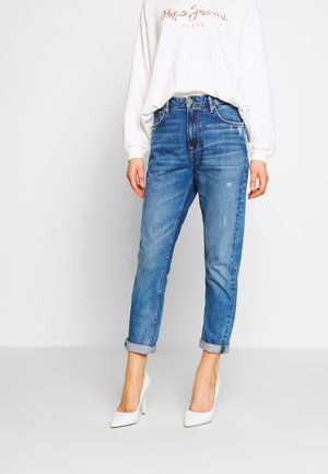 Femme portant un jean bleu retroussé, des talons hauts blancs et un sweat-shirt blanc avec un texte sur la poitrine.
