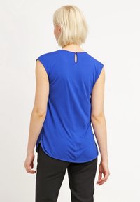 Top blu royal a maniche corte con dettaglio a oblò sul retro, orlo arrotondato e texture morbida, indossato con pantaloni neri.
