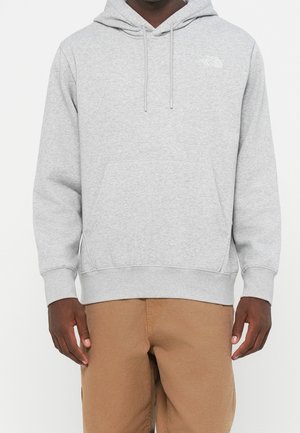 Mężczyzna ubrany w jasnoszary kaptur The North Face z przednią kieszenią i beżowe spodnie, stojący na jednolitym białym tle.