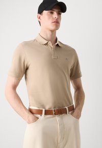 Polo beige à manches courtes, avec col et logo brodé. Associé à un pantalon crème et une ceinture en cuir marron. Casquette noire portée.