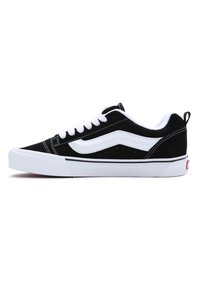 Baskets basses noir et blanc avec lacets blancs, une bande blanche ondulée sur le côté et une semelle en caoutchouc blanche sur fond blanc.