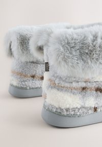 Graue Plüsch-Hausschuhe mit einem flauschigen Bündchen und einem gestreiften Muster in Creme, Grau und Hellbraun. Glatte graue Sohle an der Unterseite sichtbar.