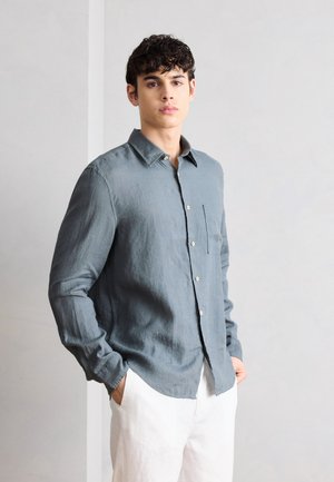 Ung mand med krøllet hår iført en grå skjorte med knapper og hvide bukser, stående med hænderne i lommerne mod en ensfarvet baggrund.