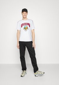 Una camiseta blanca con letras rojas que dicen "ARIZONA" y un diseño gráfico, emparejada con pantalones negros y zapatillas deportivas beige con acentos negros. Gorra negra.