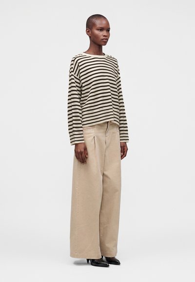 Femme portant une chemise à manches longues rayée beige et noire, un pantalon large beige et des chaussures noires pointues, debout devant un fond clair uni.
