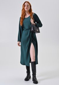 Abito lungo in satin verde con uno spacco laterale, maniche lunghe e scollo rotondo, abbinato a stivali neri al ginocchio e una borsa nera.