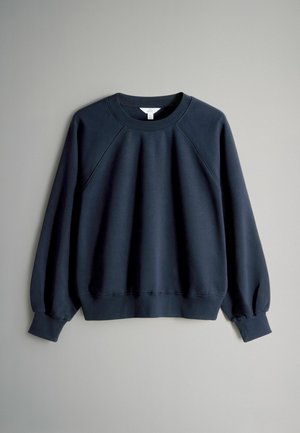 Tamsiai mėlynas džemperis su reljefiniu apykaklė ir rankogaliais, tekstūruota medžiaga ir raglaniniai rankovės. Rodykite suapvalintą hemą ir minimalų ženklinimą.