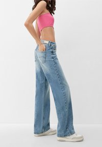 Mujer con jeans de pierna ancha azul claro, un top corto sin mangas rosa, zapatillas blancas, de pie con una mano en el bolsillo trasero.