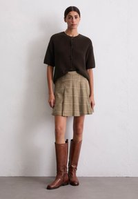 Cardigan court marron à côtes, jupe beige à carreaux et bottes en cuir marron haute. La jupe présente des plis et un motif texturé.