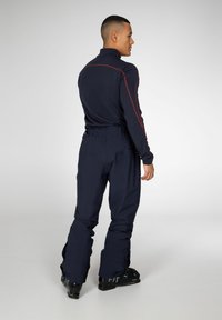 Marineblauwe thermoshirt met hoge kraag en rode stiksels, gecombineerd met marineblauwe skibroeken. De broek heeft een taille-sluiting en wijde broekspijpen. Zwarte skibotten.