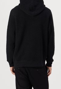 Schwarzer Fleece-Hoodie mit strukturiertem Äußeren, verstellbare Kapuze und gerippten Bündchen. Kombiniert mit schwarzen Hosen mit Knopf-Akzenten.