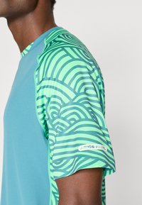 Αθλητική μπλούζα κοντομάνικη με υφασμάτινο μπλε σώμα και πράσινα μανίκια με κυκλικές ρίγες. Λογότυπο "ASICS PADEL" στο παντελόνι.