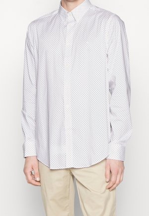 Wit, lang mouw, overhemd met knopen en een kleine donkere geometrische print, gedragen door een persoon met beige broek tegen een effen achtergrond.