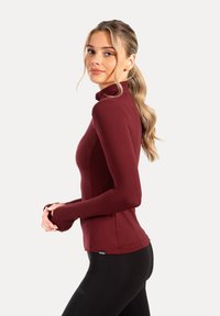 Bourgogne langærmet bluse med høj krave, glat tekstur, tætsiddende design og tommelhuller, parret med sorte leggings.